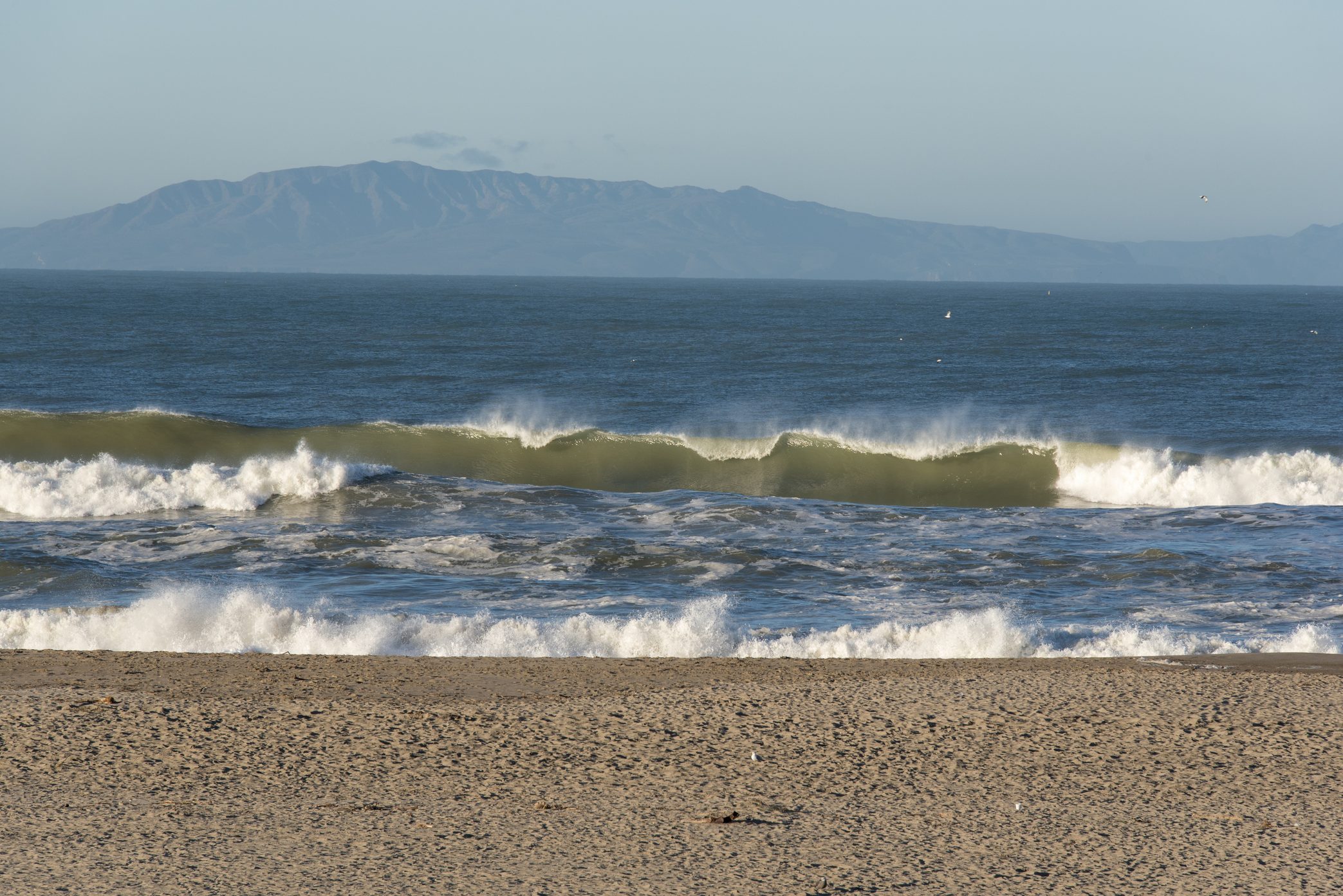 Waves crash on a sandy beach, with a mountain range visible in the background under a clear blue sky.