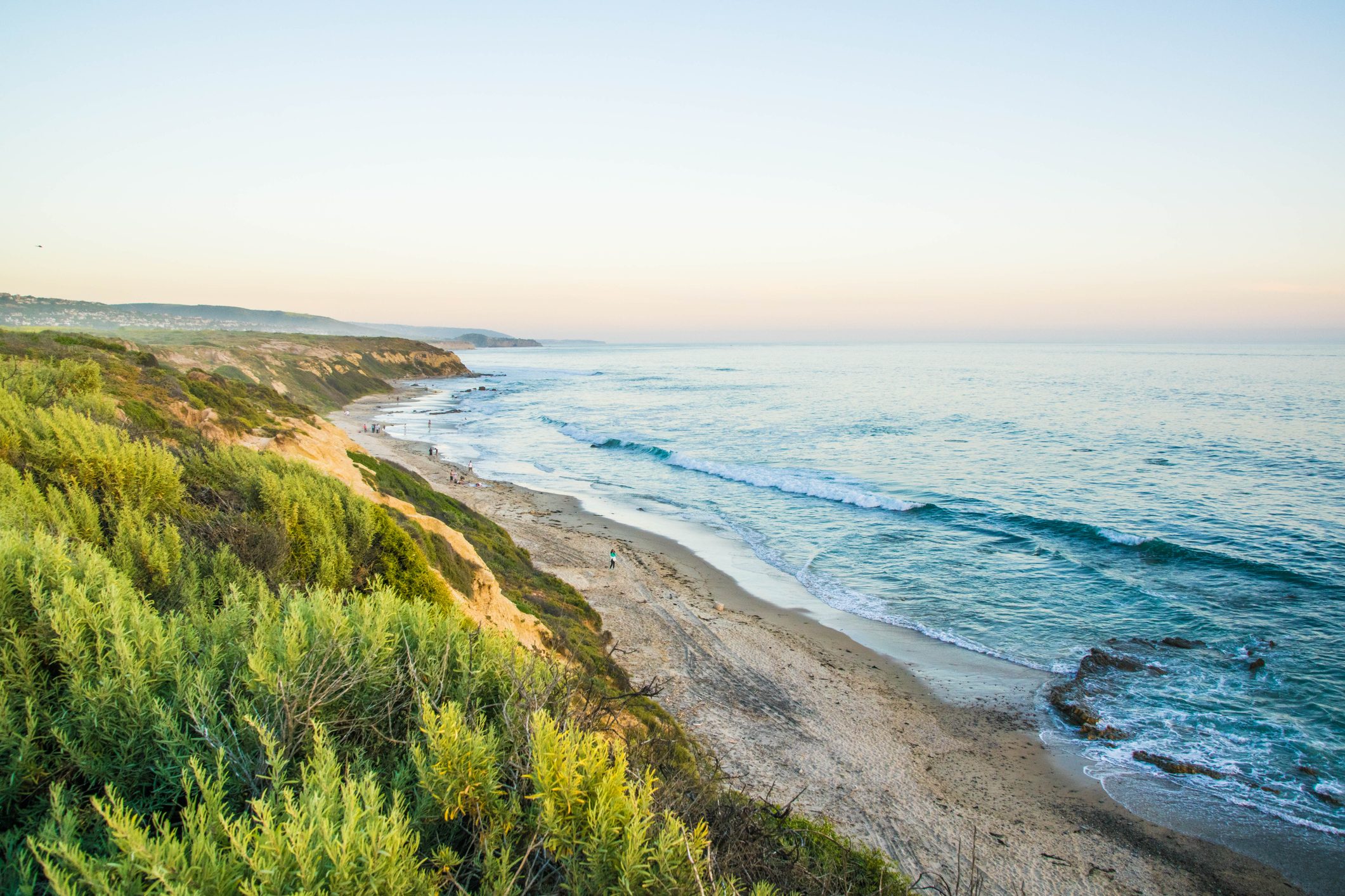 Waves crash gently on a sandy beach bordered by green cliffs, with people strolling along the shoreline under a clear sky.
