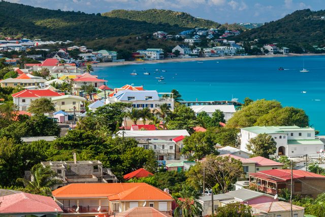Colorful houses sit quietly along the coast, surrounded by lush greenery, overlooking a turquoise bay with boats. Hills rise in the background.