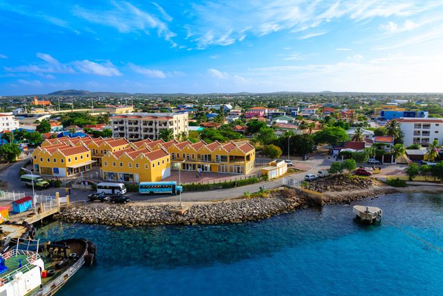 Yellow buildings line a coastal road with parked vehicles; a vibrant townscape and clear blue sky provide the backdrop.