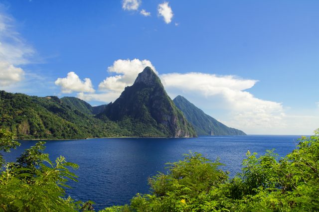 Mountain rises dramatically, covered in lush green vegetation, beside a serene blue ocean under a clear sky with scattered clouds.