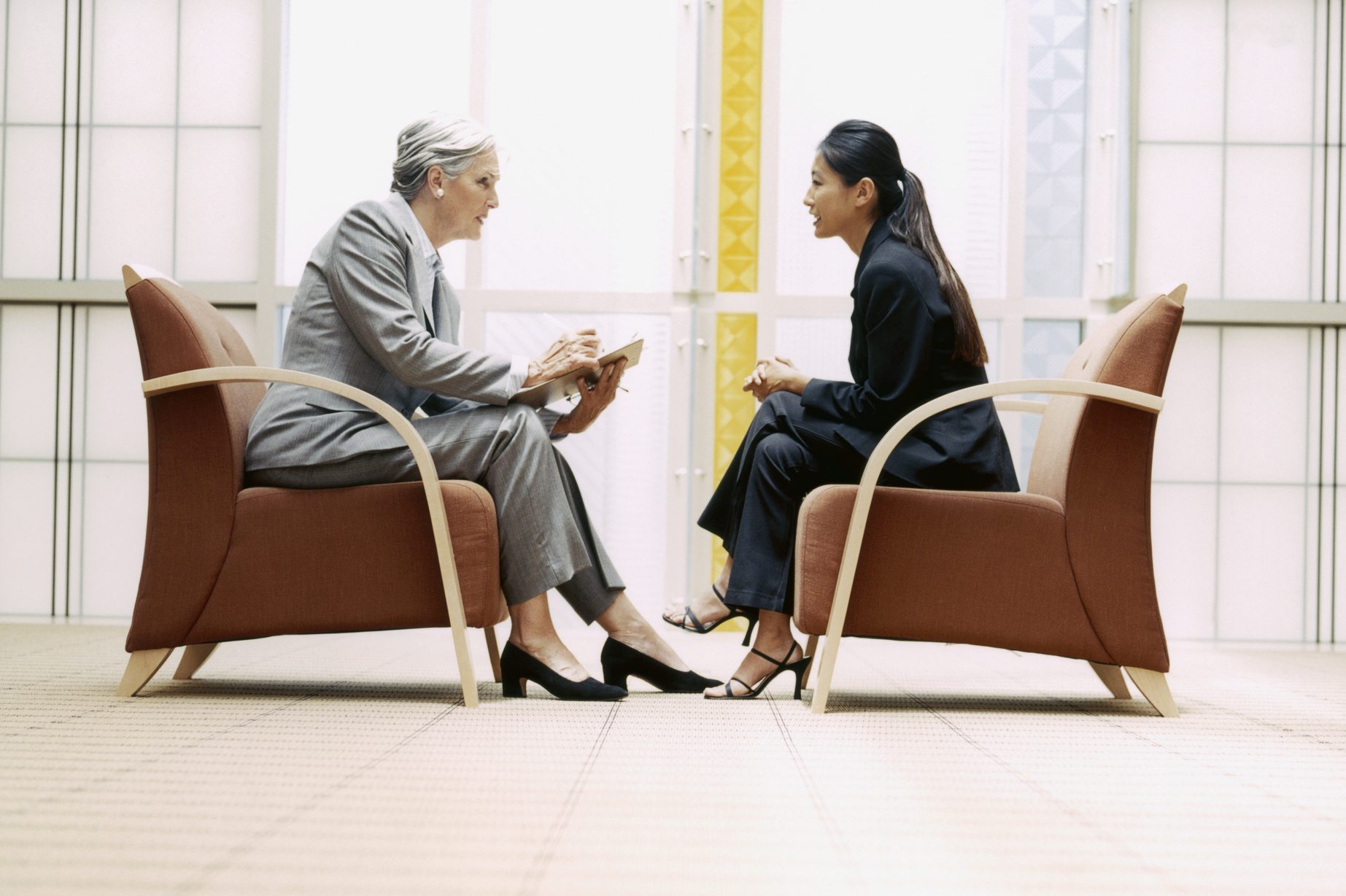 Two people sit in armchairs, engaged in a conversation, in a modern, bright office setting.