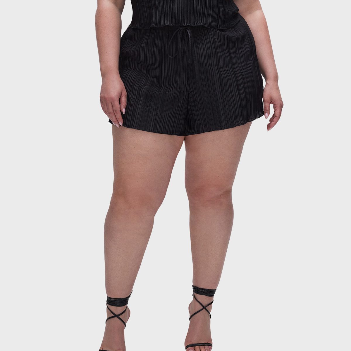 A person stands wearing a black pleated romper with drawstring waist, paired with strappy sandals; plain white background.