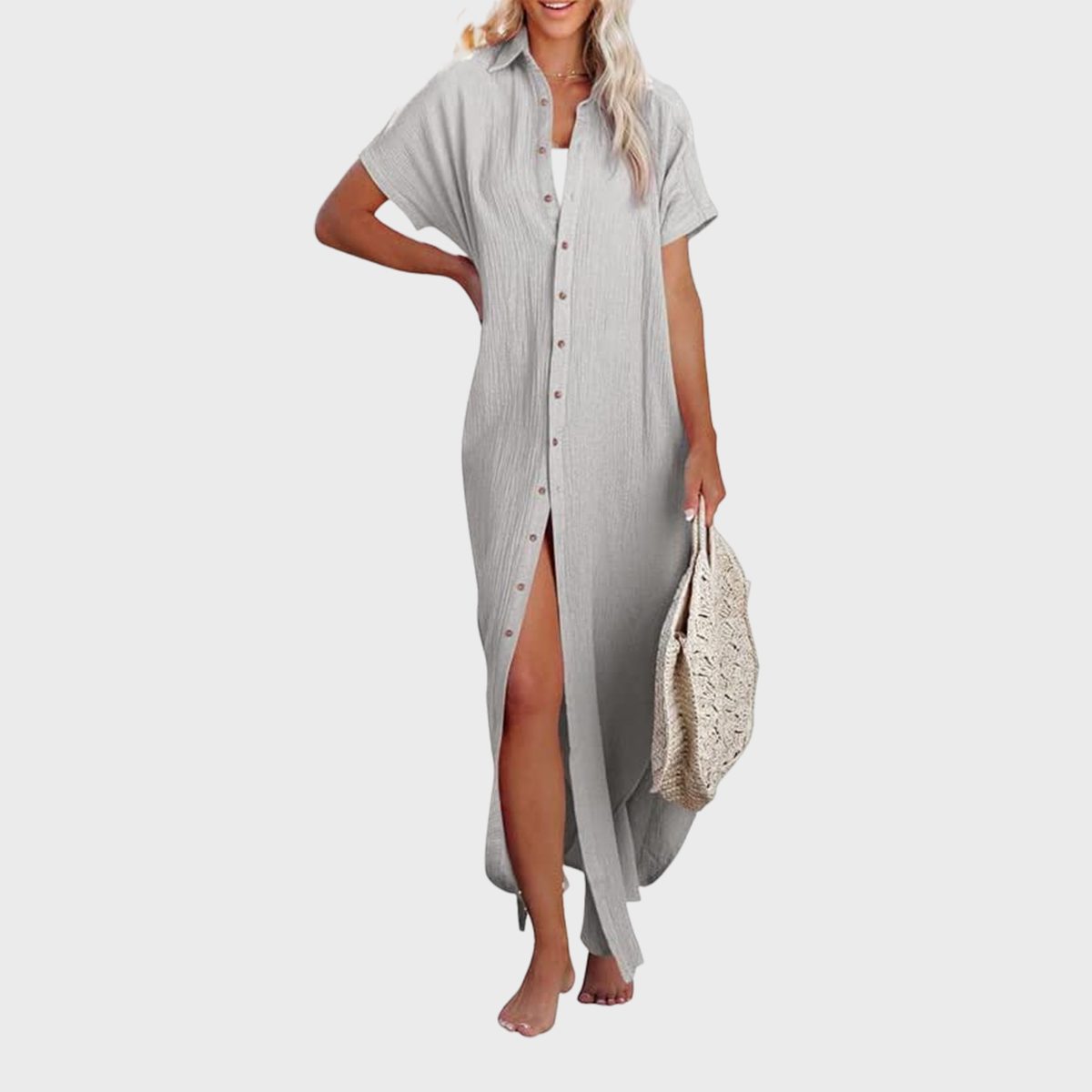 A person wears a long, light-gray button-up dress, holding a woven bag, standing barefoot against a plain white background.