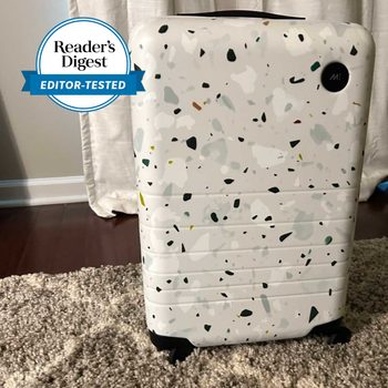 Speckled suitcase stands on a beige carpeted floor in front of curtains. A Reader's Digest "Editor-Tested" badge is displayed.