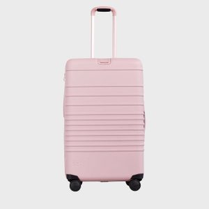 Pink suitcase stands upright, handle extended, on a smooth surface; features four wheels and 