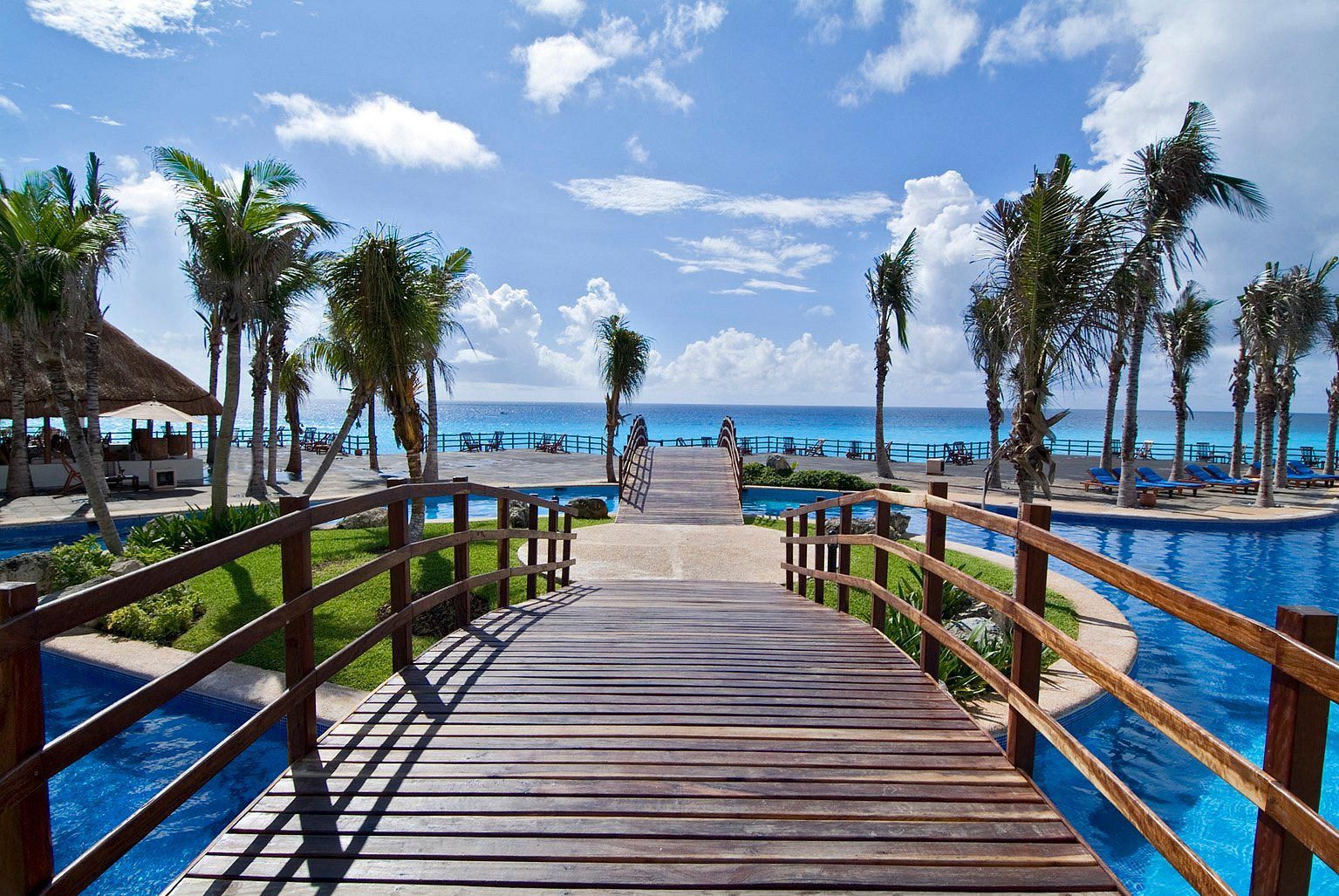 A wooden bridge spans a pool area, surrounded by palm trees, leading towards the ocean under a bright, blue sky.