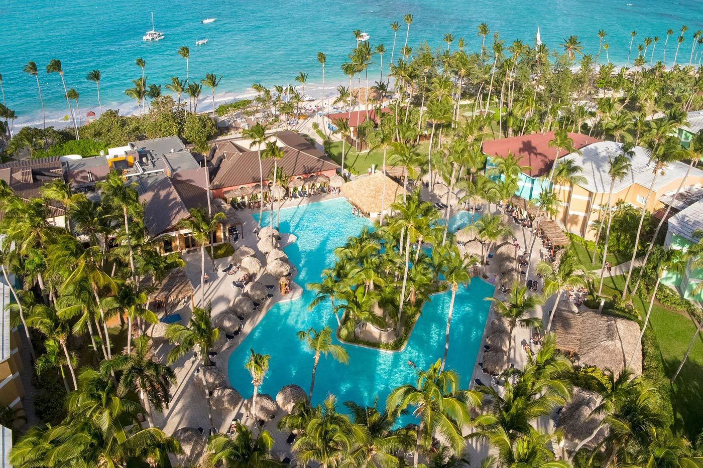 Swimming pool surrounded by palm trees and thatched-roof huts, situated near a beach with turquoise waters and boats on the horizon.