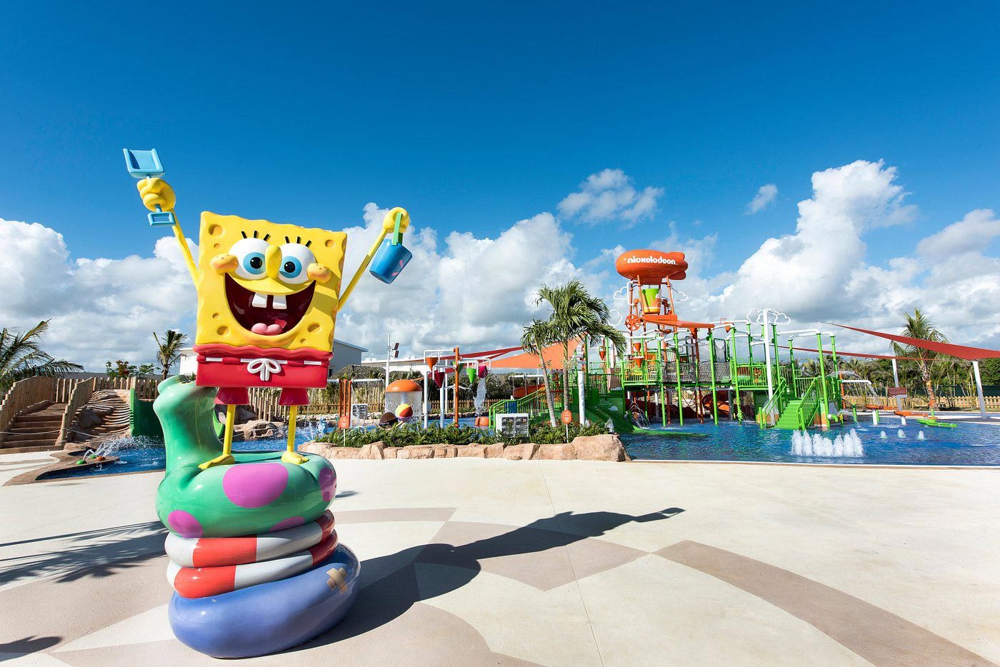 A SpongeBob statue stands joyfully on inflatables at a colorful water park, under a bright blue sky with scattered clouds.