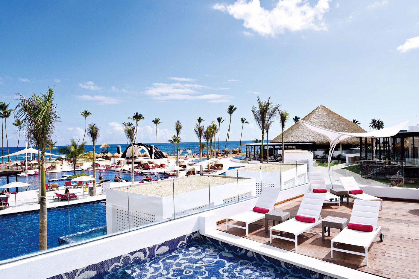 Lounge chairs rest poolside with ocean view; surrounding are palm trees, a thatched-roof structure, and sunny sky, creating a tropical resort atmosphere.