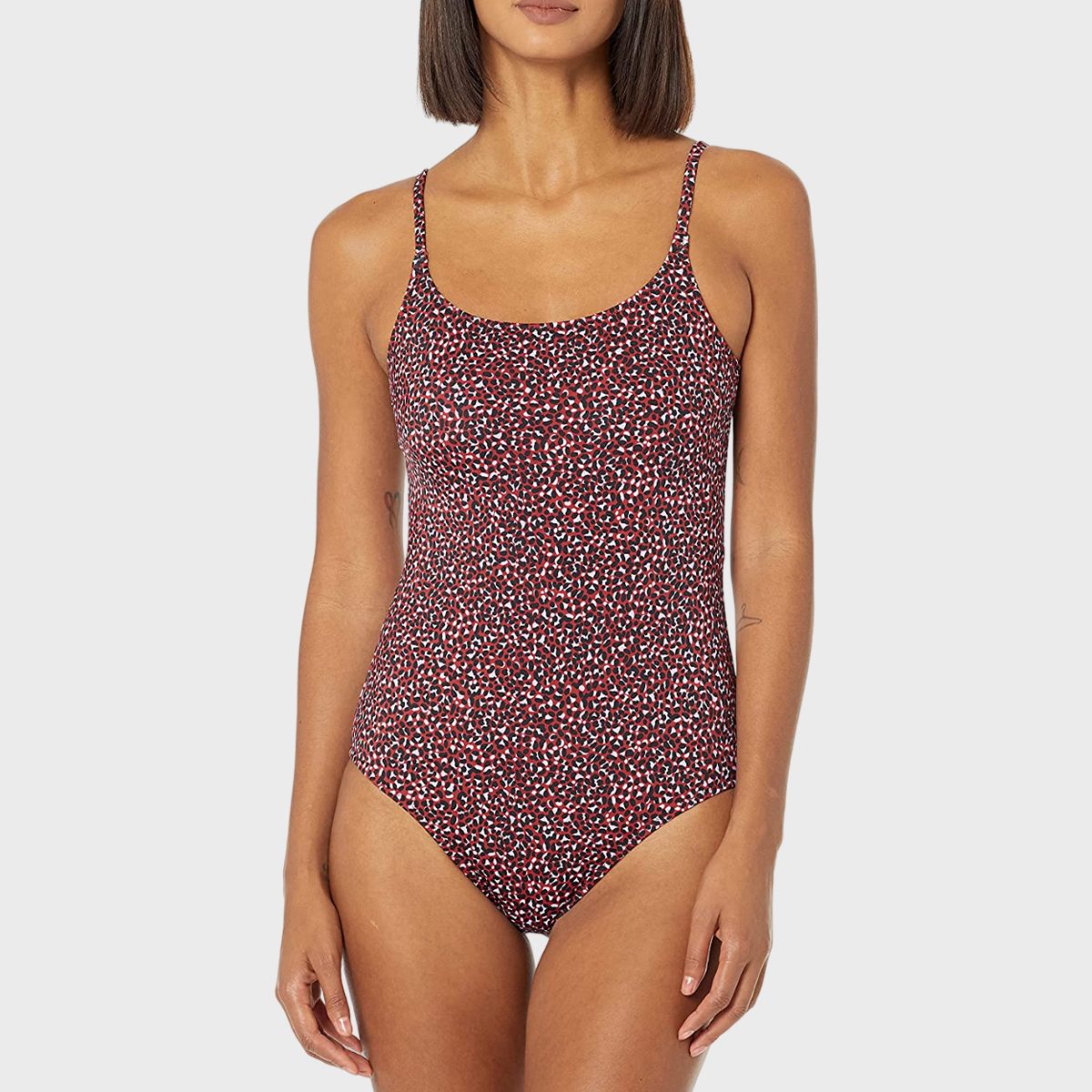 A woman stands modeling a red and black patterned one-piece swimsuit against a plain gray background.