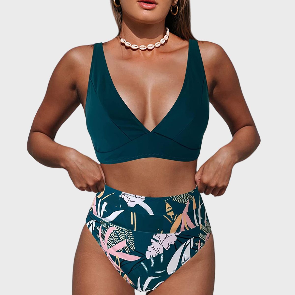 Woman stands adjusting high-waisted floral bikini bottoms, wearing a teal top, backdrop of plain white.