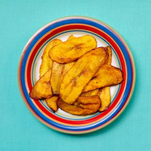 Fried plantain slices rest on a colorful, striped plate set against a teal background.