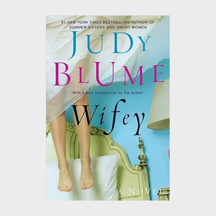 Feet dangle above bed, covered by a sheet, in a brightly colored bedroom; "Judy Blume Wifey" text prominently displayed in the foreground.