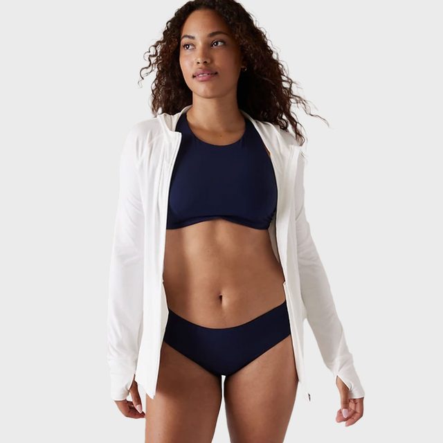 Woman wearing a navy swimsuit and white jacket stands confidently against a plain, light gray background.