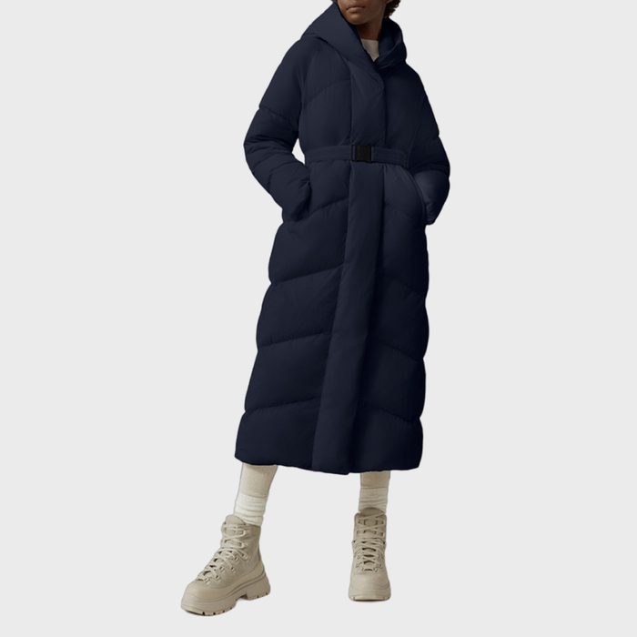 A person wears a long, dark puffer coat with a belt, standing on a plain, light background. They have beige boots and white socks.