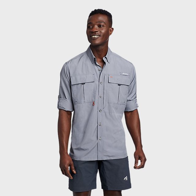 Man smiles, wearing a gray collared shirt with rolled sleeves and dark shorts, standing against a plain light gray background.