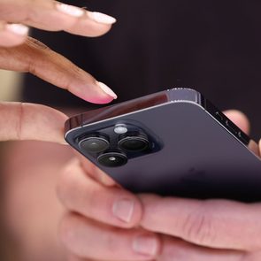 Hands hold a smartphone as a finger points at its camera lenses in a close-up, indoor setting.
