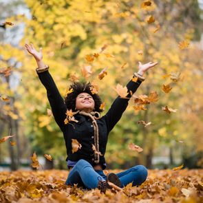 Person sits joyfully, tossing autumn leaves, surrounded by a colorful forest.
