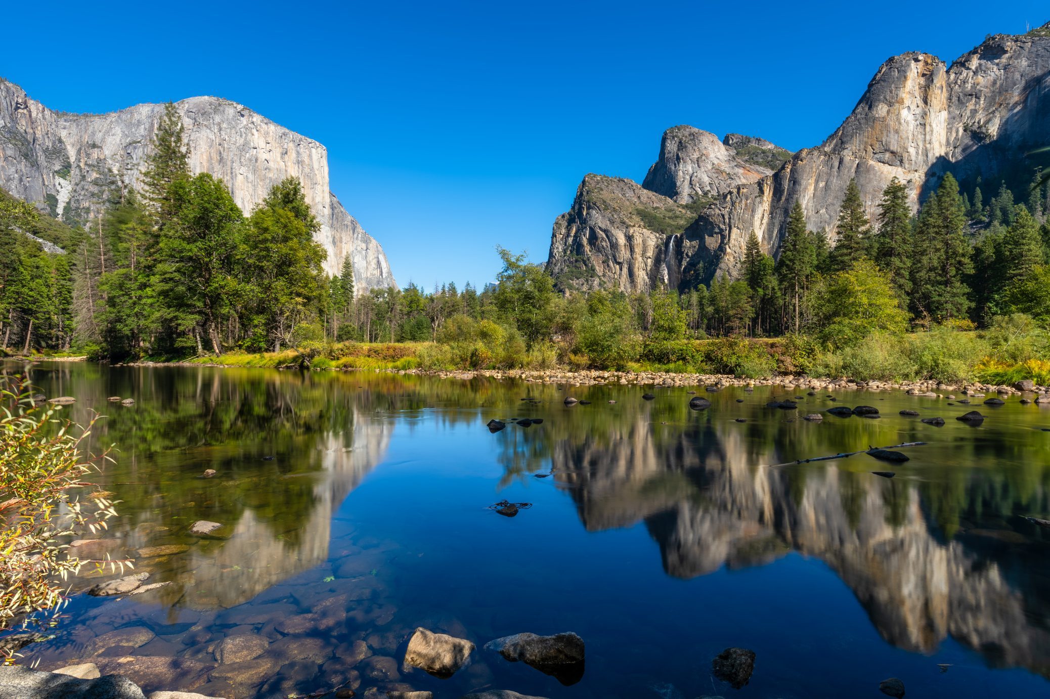 Mountains reflect in a calm river surrounded by lush green trees under a clear blue sky.