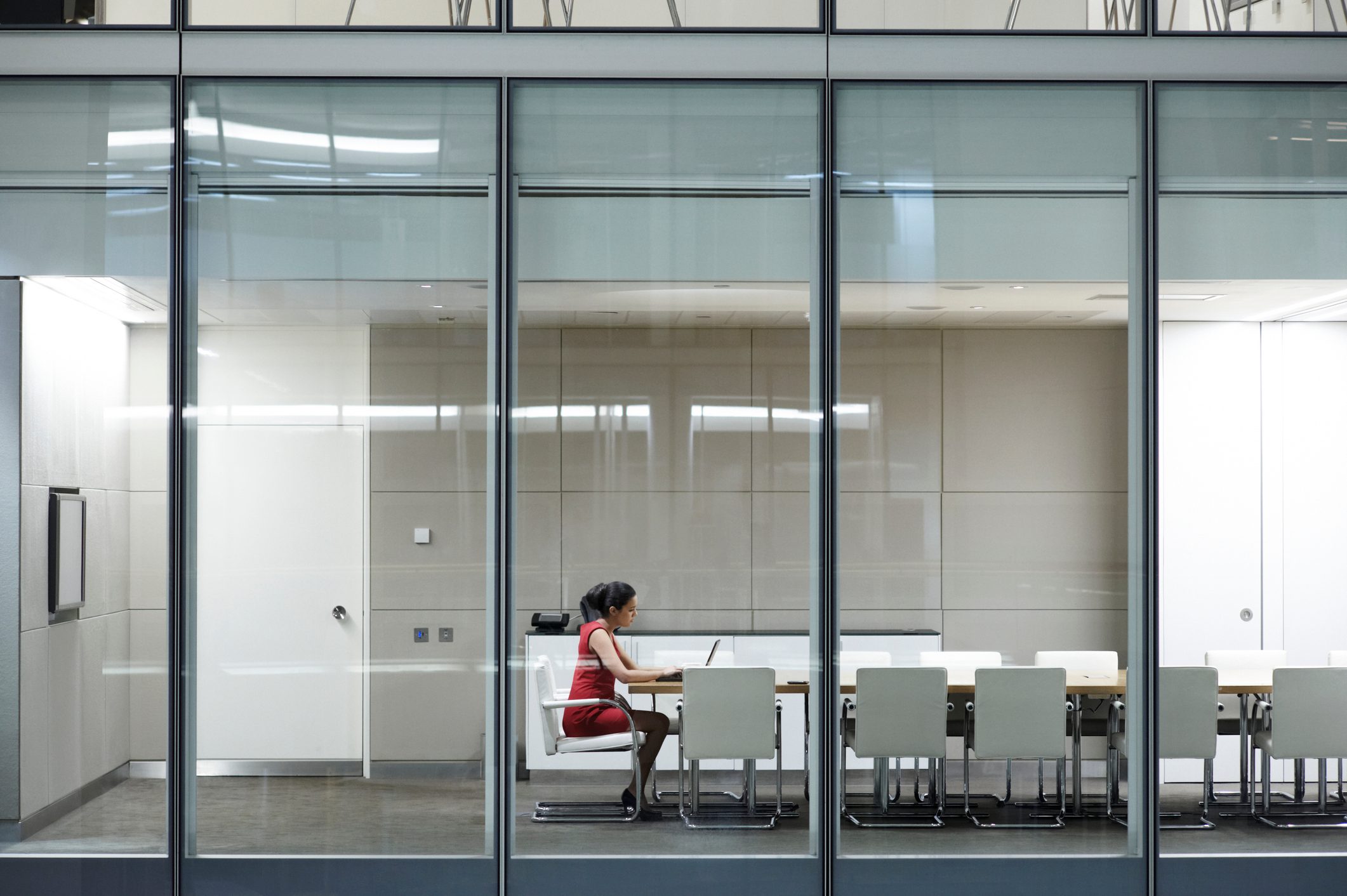 A person types on a laptop inside a glass-walled conference room, surrounded by empty chairs and modern office furnishings.