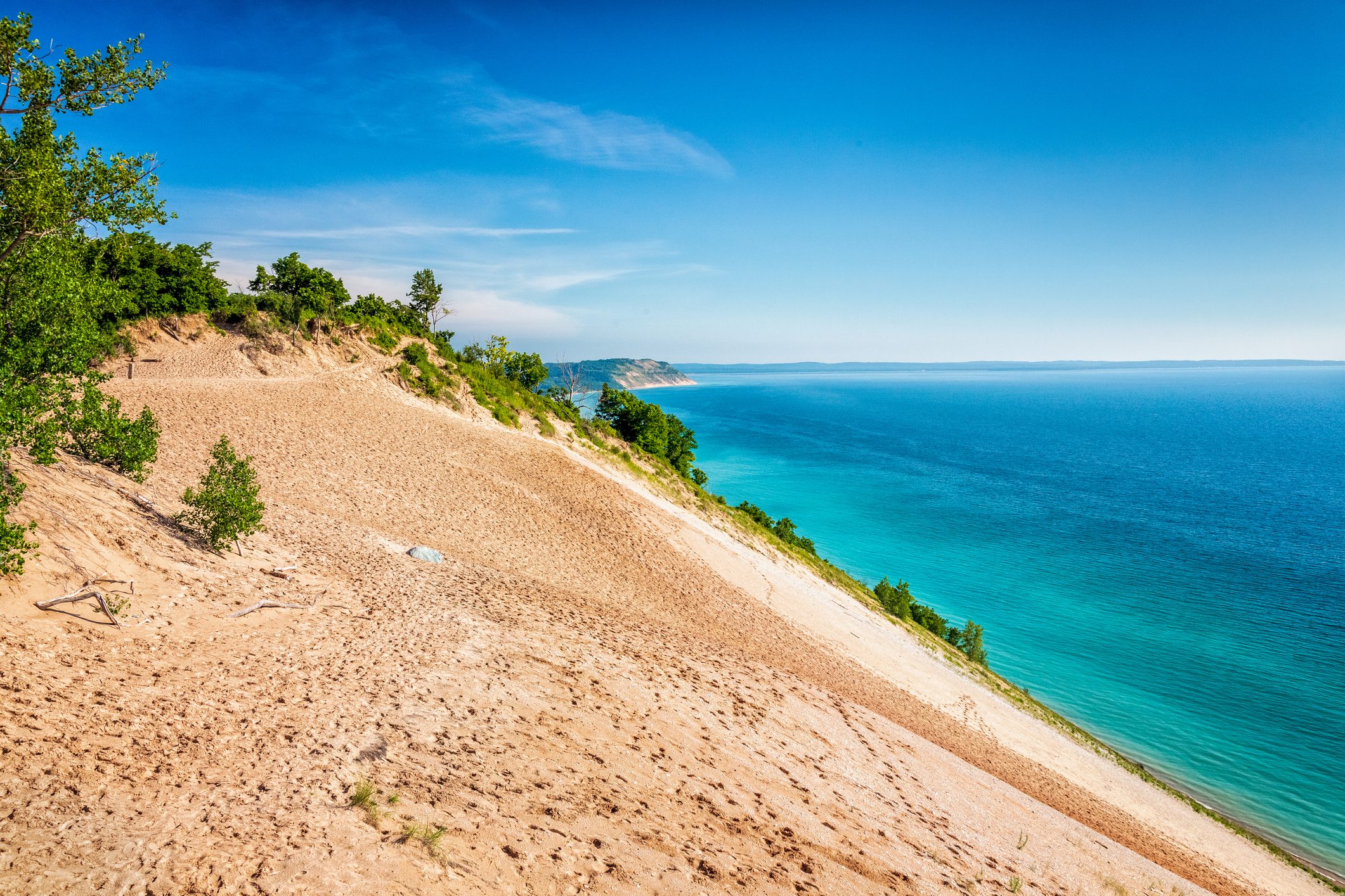 Sandy dune slopes toward a vast, turquoise lake, bordered by green vegetation under a clear blue sky.