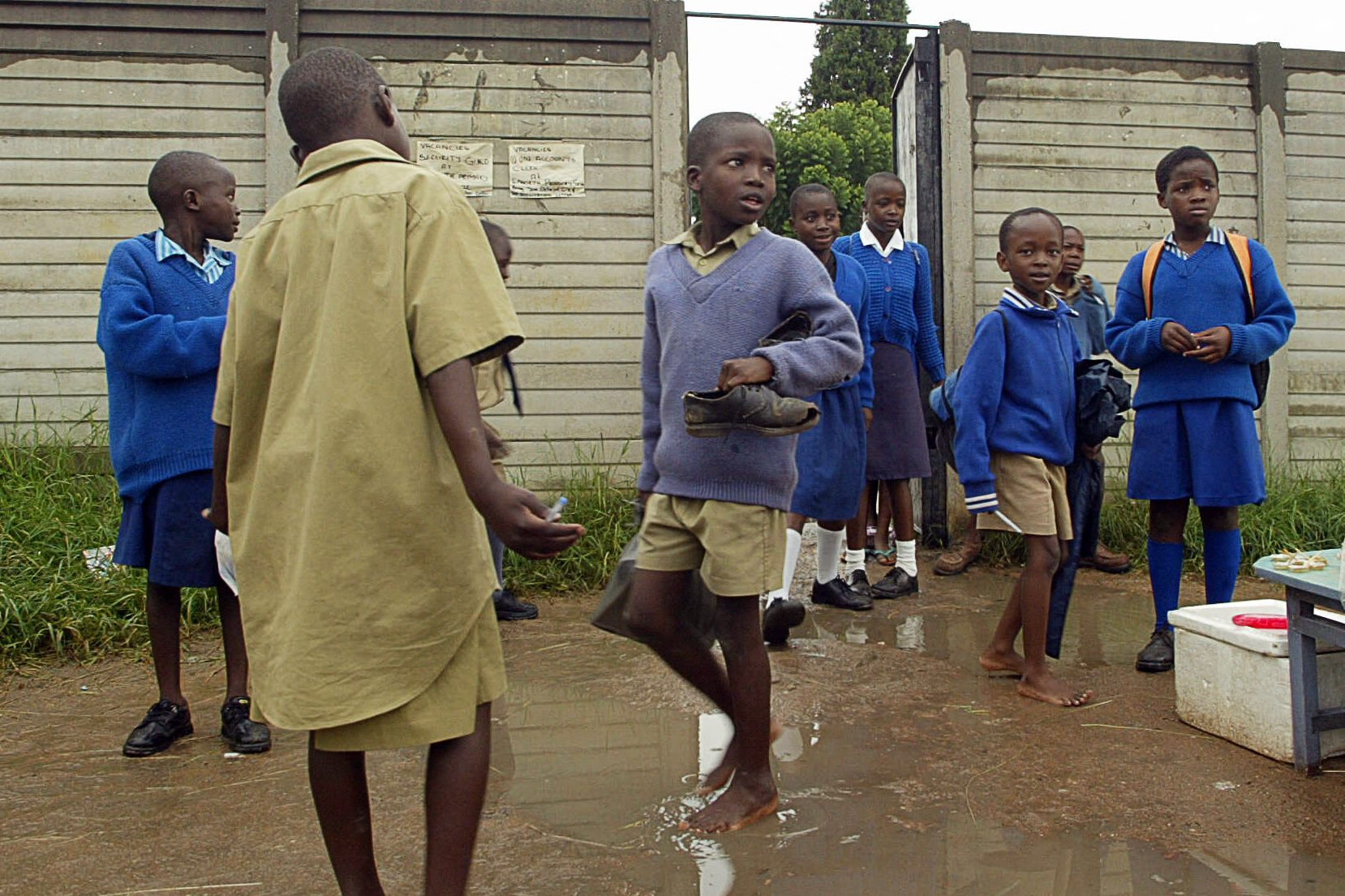 Children, in school uniforms, navigate a muddy puddle beside a concrete wall with posted signs in an outdoor setting.
