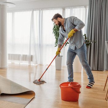 A person mops the wooden floor in a bright, modern living room near a couch and bar stools.