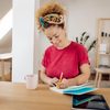 A woman is writing in a notebook at a wooden table, surrounded by a plant, mug, and tablet in a cozy living space.