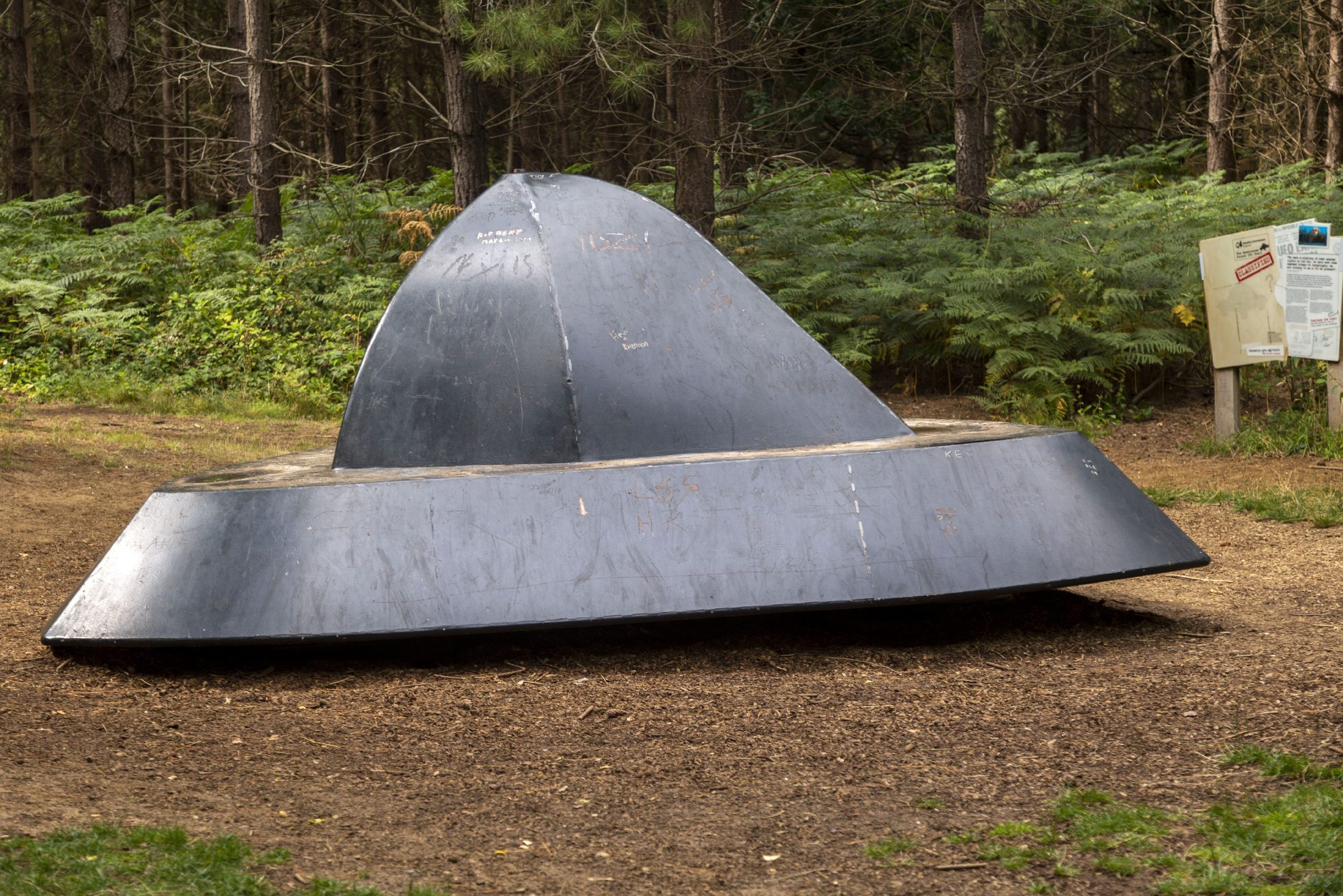 Metal UFO sculpture rests on forest ground near informational sign amidst dense greenery and trees.