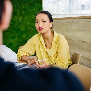 Person in yellow shirt sits at table, holding paper, engaged in conversation. Indoor setting with green textured wall and wooden paneling.