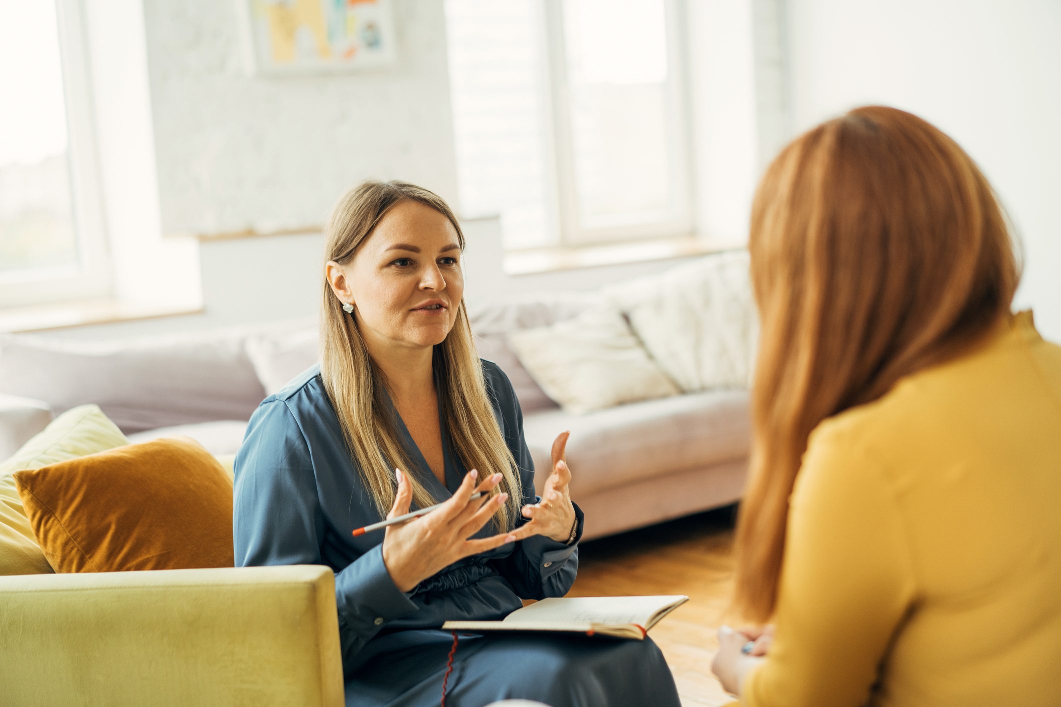 A woman in blue gesticulates while holding a notebook and pen, conversing with another person in a cozy living room.