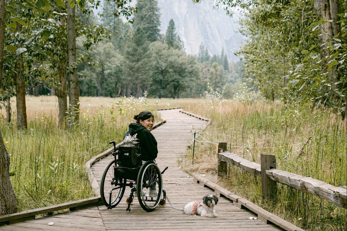 Person in wheelchair smiling on wooden path, next to small dog, surrounded by lush forest and grassy field.