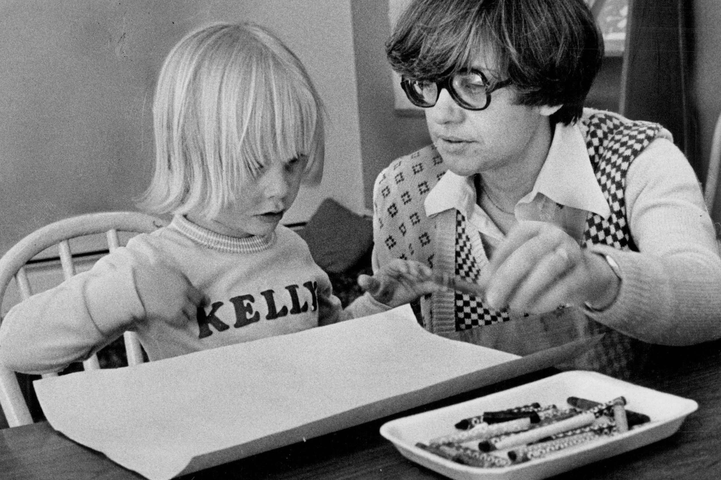 Child drawing with crayons, adult assisting, at a table. Shirt reads "Kelly," crayons lie on a nearby plate. Indoor setting, focused concentration.