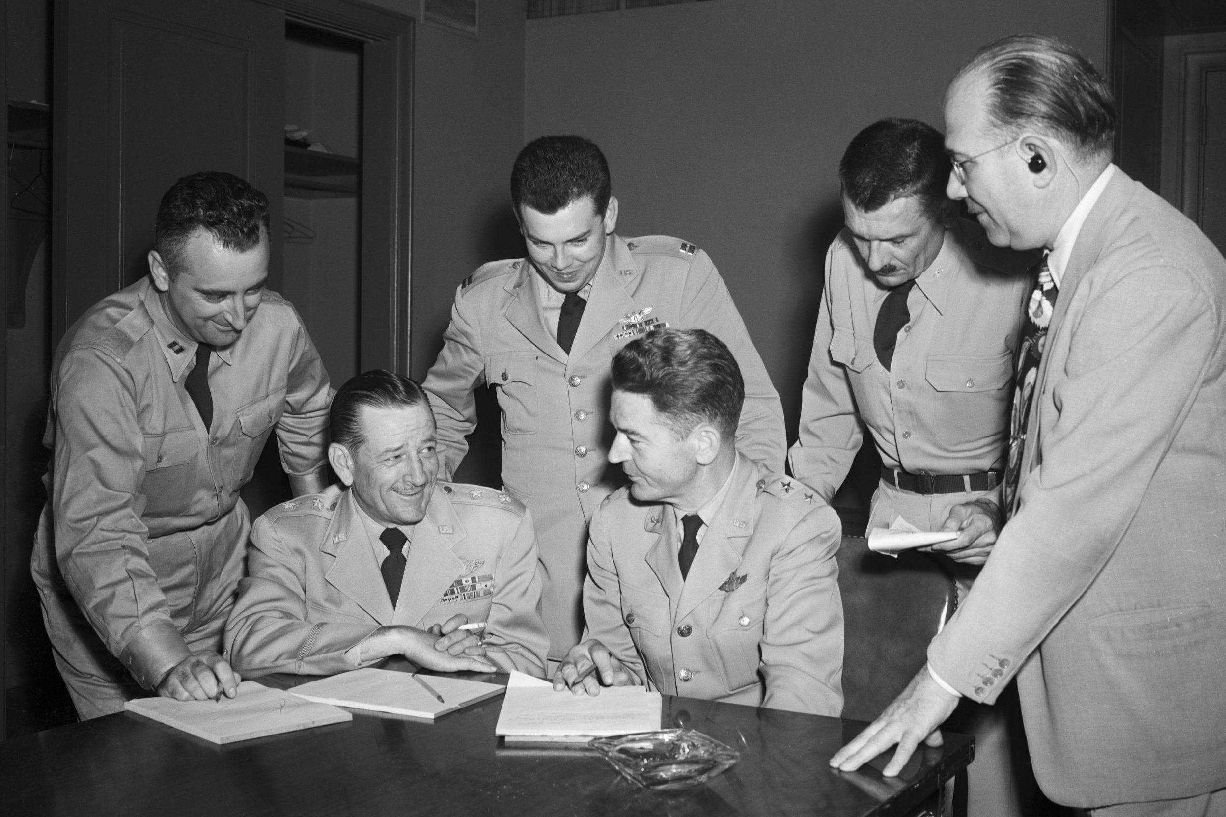 Six men in military uniforms and suits discuss papers at a table, in an office setting.