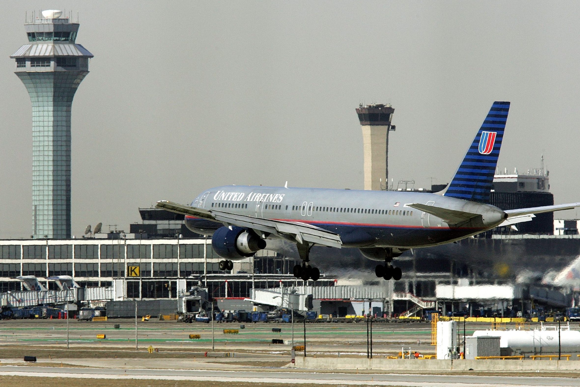 Aircraft landing on a runway, surrounded by airport buildings and control towers, with "United Airlines" written on the fuselage.