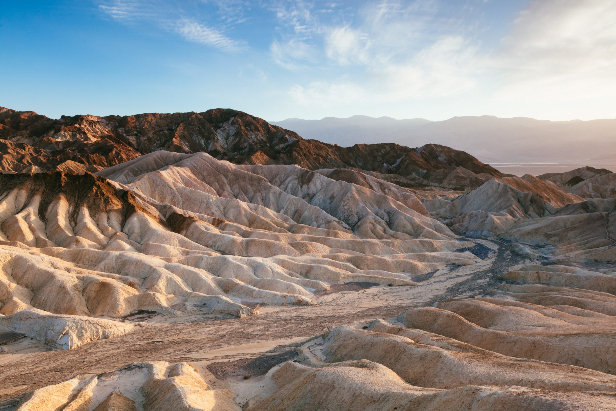 Eroded hills span the landscape, illuminated by soft sunlight, under a clear blue sky, creating a rugged and desolate desert scene.