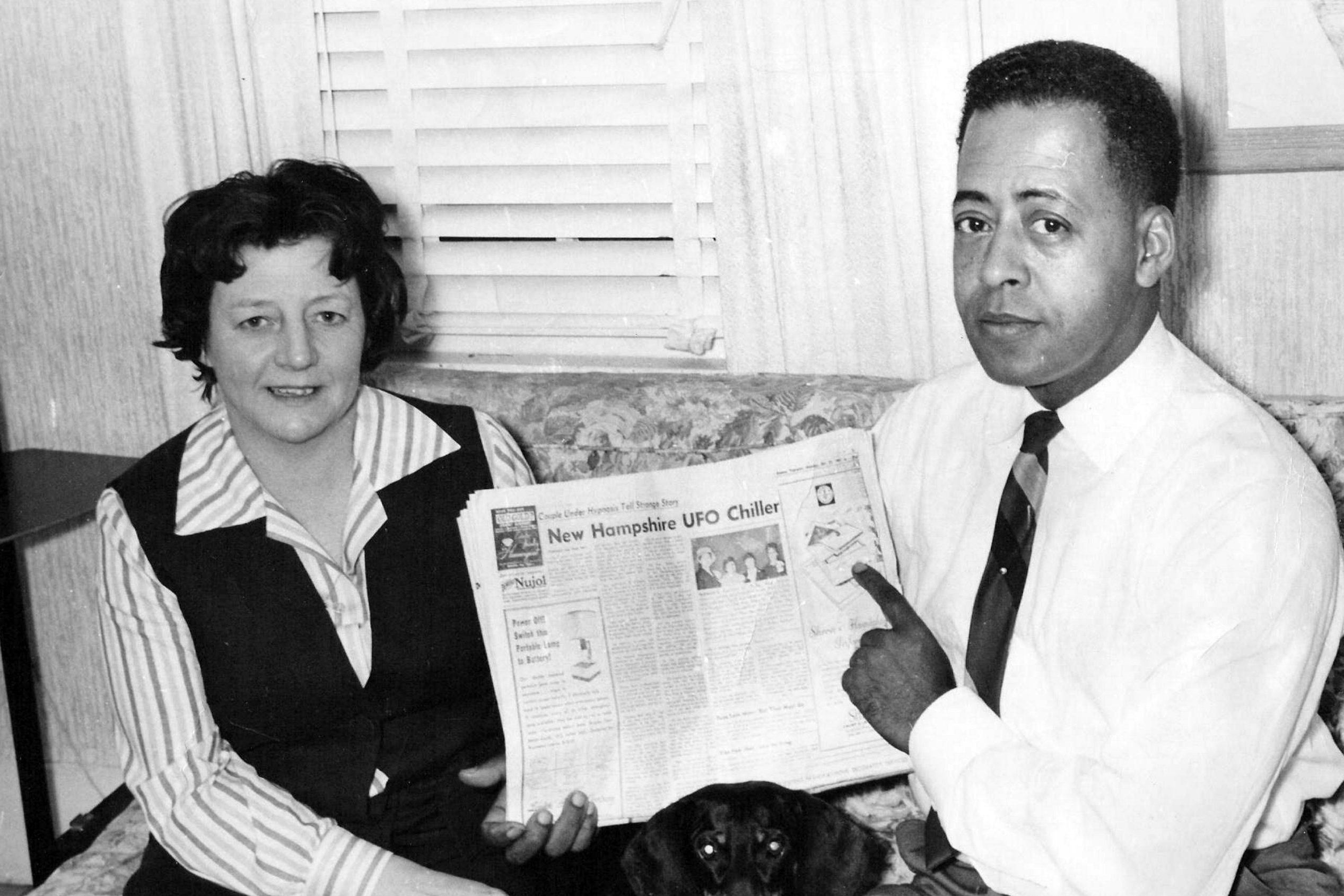 A person points to a newspaper featuring a "New Hampshire UFO Chiller" headline; another sits beside them on a couch with blinds behind.
