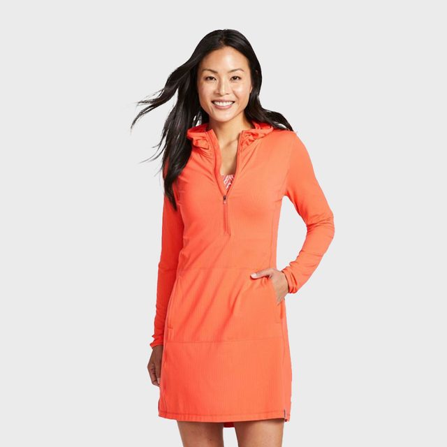 A woman wearing an orange sporty dress smiles with one hand in pocket against a plain gray background.