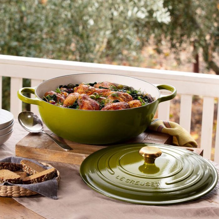 Green pot holds cooked meal on wooden table, outdoors with bread and plates nearby; â€œLe Creusetâ€ on lid in foreground.