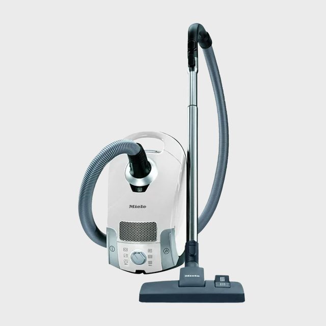 Vacuum cleaner stands upright; hose attached to base; gray context suggests a modern, sterile setting. The vacuum is labeled with 