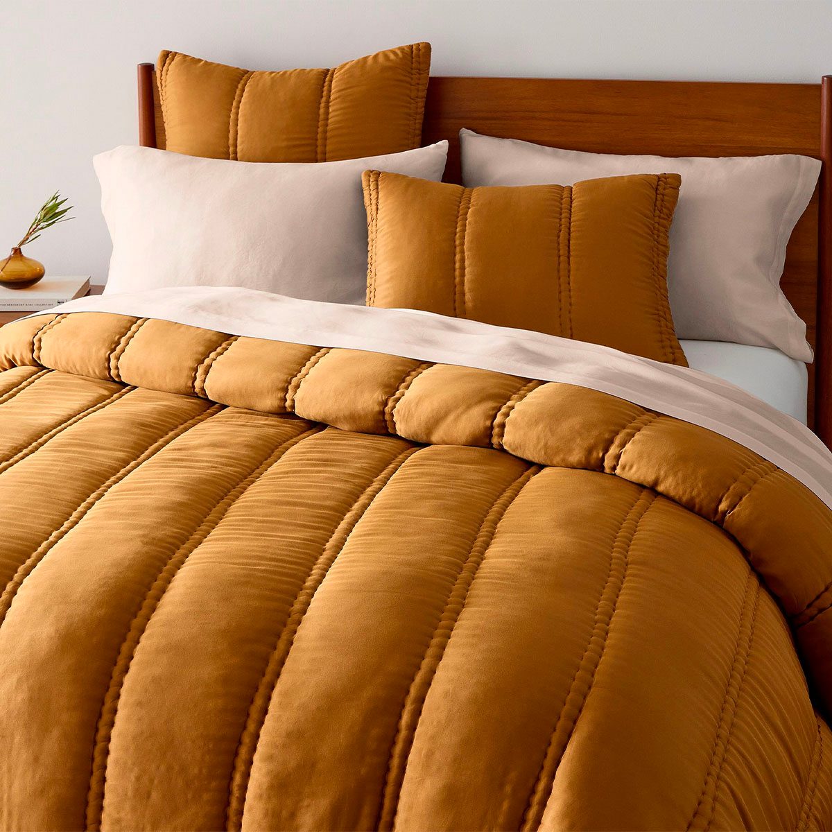 A mustard-colored bedspread neatly covers a bed with matching pillows and a wooden headboard; a small vase sits on a bedside table.