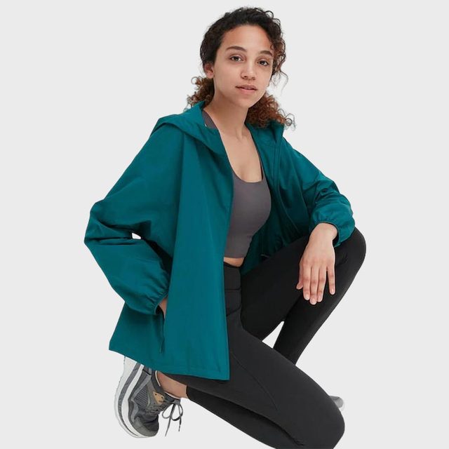 Person squats, wearing teal jacket, gray top, black leggings, and sneakers in a neutral environment.