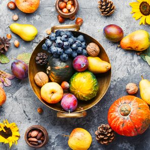 Bowl holding assorted fruits and nuts, surrounded by pumpkins, pears, apples, plums, sunflowers, pinecones, and leaves on a textured gray surface.