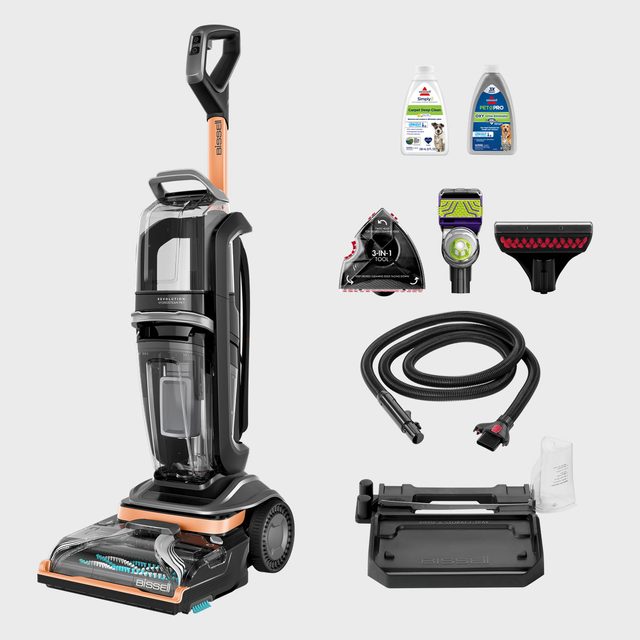 Vacuum cleaner stands upright, surrounded by various attachments and two bottles of cleaning solution against a plain background.