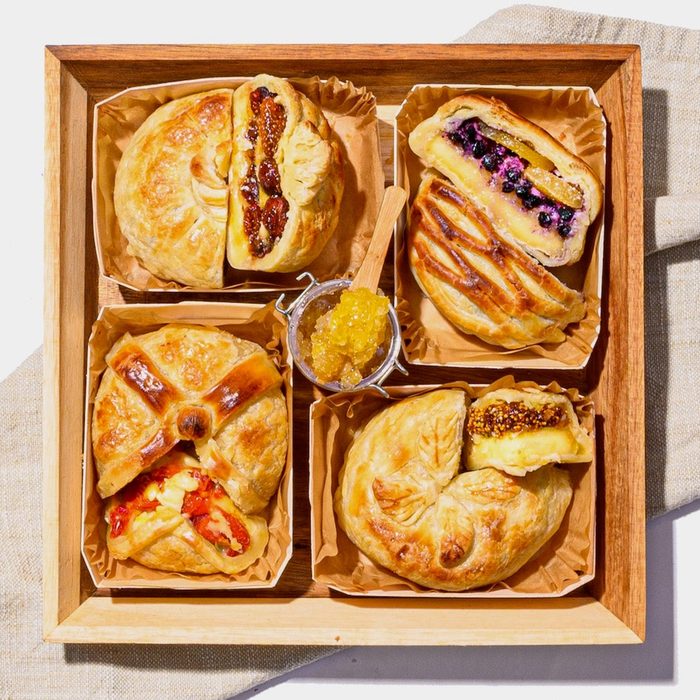 Pastries rest together in a wooden tray, each filled with varied ingredients like fruit and cheese, with a small jar of yellow jam.