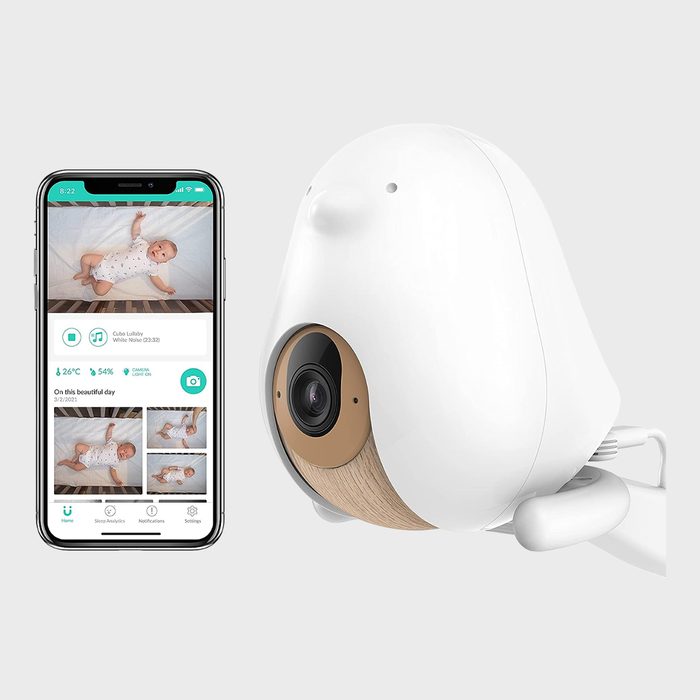 A white baby monitor camera displays images on a smartphone screen, showing baby photos and app interface in a minimalistic setting.