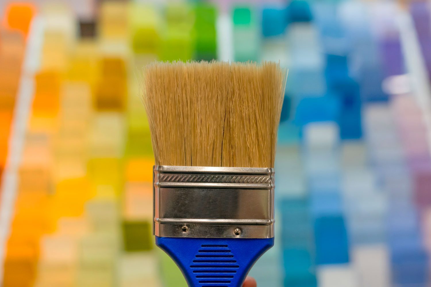 A paintbrush is held upright against a blurred background of colorful paint swatches arranged in a gradient from warm to cool tones.