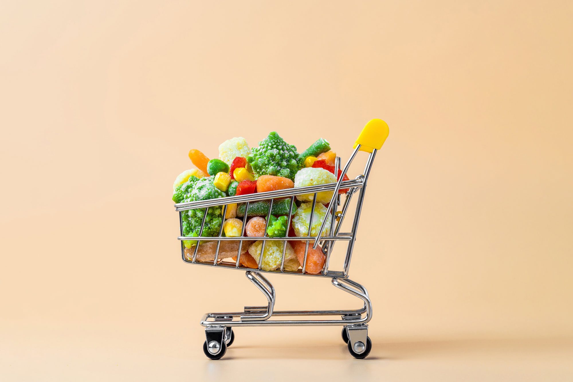 Shopping cart holds frozen vegetables against a plain, light peach background.