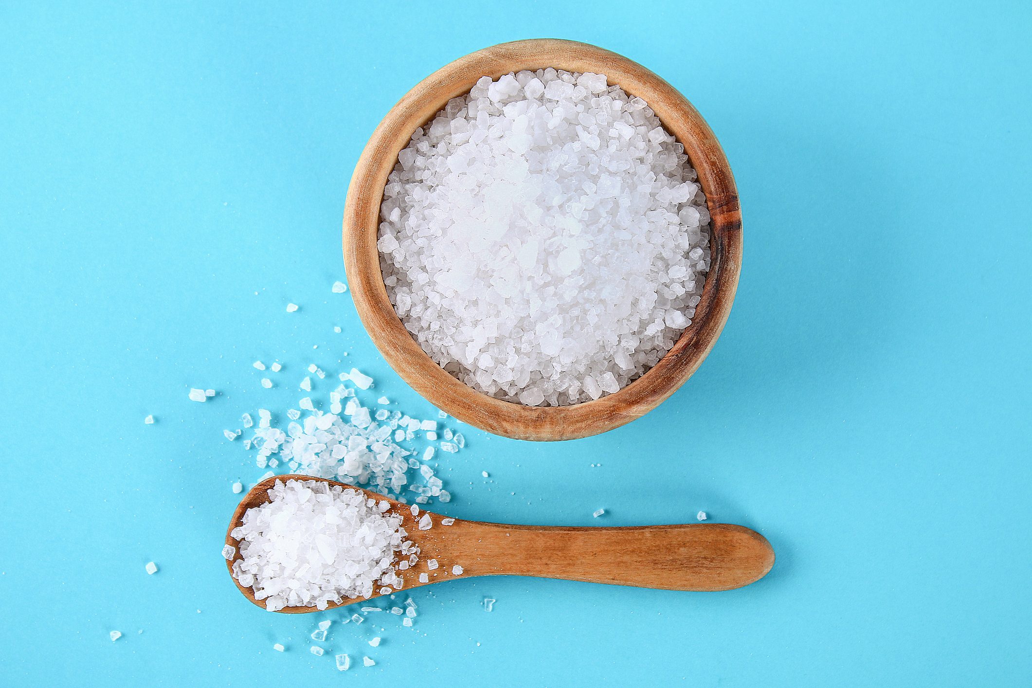 Salt crystals fill a wooden bowl and spoon, resting on a blue surface. Some crystals are scattered around.
